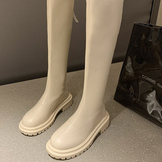 Paire de bottes haute grise avec semelle caoutchouc, fond gris avec un sac noir au second plan.