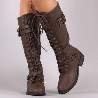 Paire de bottes à lacets marron sur fond gris et portées par un individu dont on voit que les mollets.