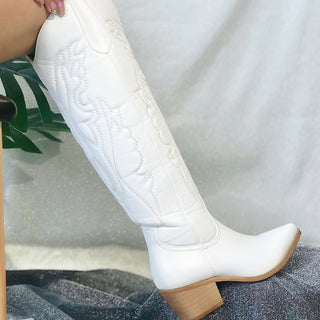 Botte de cowboy blanche portée avec un talon en bois posée sur un tissu gris avec une plante verte derrière