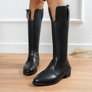 Photo d'une paire de botte cavalière noir en cuir PU lisse fendu au dessous du genoux, portées sur d parquets devant un mur blanc.