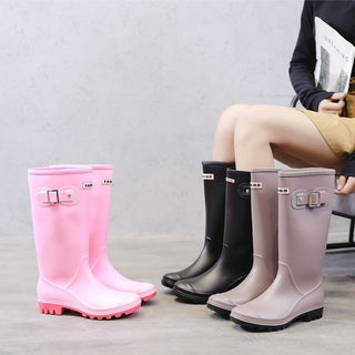 Buste de femme assise portant des chaussures de pluie noires avec d'autres chaussures roses et gris posées à côté.