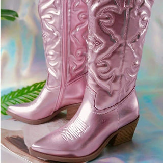Paire de bottes roses style santiag effet métallisé, avec semelle et talon marron. De trois-quarts les deux. Botte droite vue de l'intérieur avec la fermeture éclair, botte de gauche vue de l'extérieur. Sur fond papier argenté.