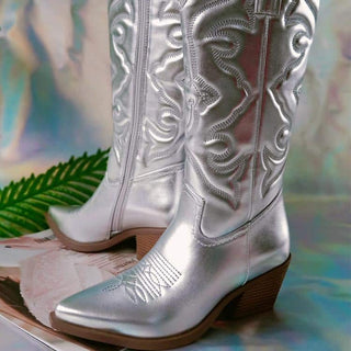 Paire de bottes style cowboy grise effet métallisé, avec semelle et talon marron. De trois-quarts les deux. Botte droite vue de l'intérieur avec la fermeture éclair, botte de gauche vue de l'extérieur. Sur fond papier argenté.