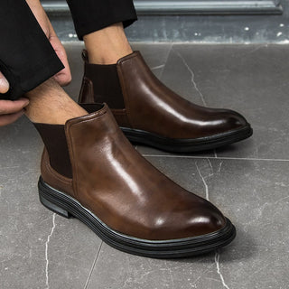 Paire de bottines homme courte marron à élastique, la semelle est noire et le bout de la chaussure est plus foncée. Portées par une mannequin, on ne voit que ses bas de jambes et le bas de son pantalon, ainsi que quelques doigts qui retroussent le pantalon. Sur un sol gris.