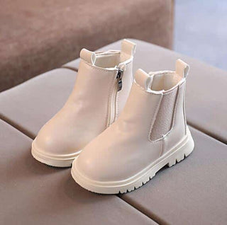 On voit de très jolies petites bottines couleur crème pour petite fille. Elles sont posées sur un canapé gris.
