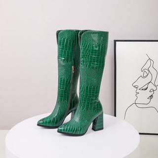 Paire de bottes à talons vertes en imitation peau de serpent posées sur une table ronde blanche avec un cadre derrière avec un dessin noir sur blanc à l'interieur
